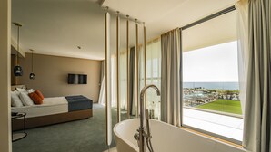 A modern hotel room with a large window, a bathtub, a bed, and a TV.