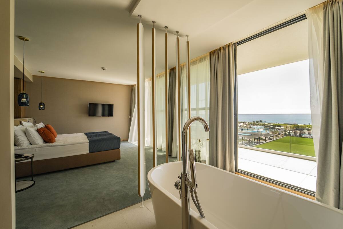 A modern hotel room with a large window, a bathtub, a bed, and a TV.