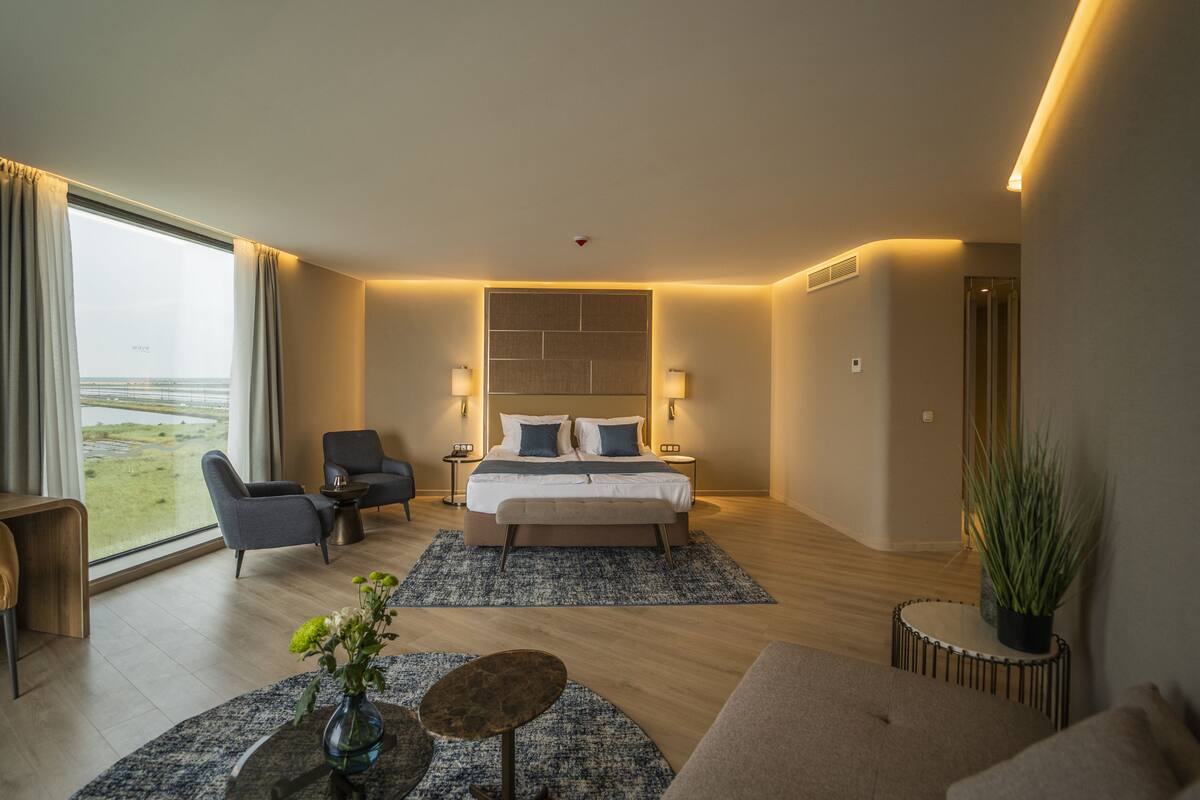 A modern hotel room with a large bed, two bedside tables, a sitting area with a sofa and armchairs, a small table with a vase, and a view of a body of water through the window.