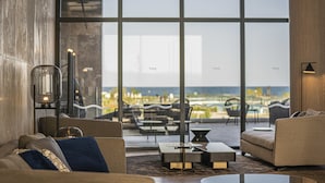 A modern living room with a large glass wall, a sofa, a coffee table, and a floor lamp. - Wave Resort – Ultra All Inclusive (Pomorie)