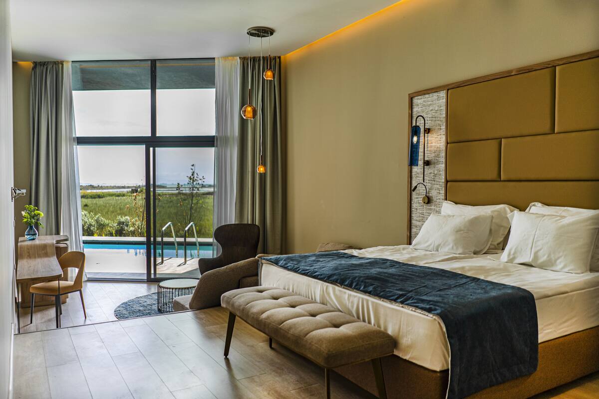 A hotel room with a large bed, a bench, a chair, a small table, and a view of a swimming pool.