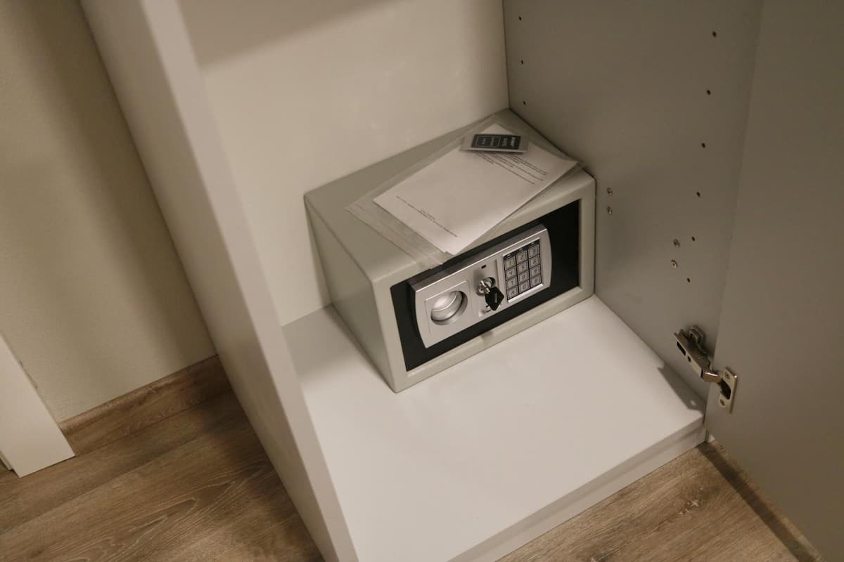 A small, white, metal-framed safe with a keypad and a remote control on top, placed on a white shelf against a wall.