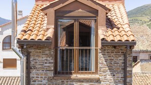 A stone building with a tiled roof, a large window with a metal railing, and a chimney. - Casas Rurales Abuela Benita (Cebreros)