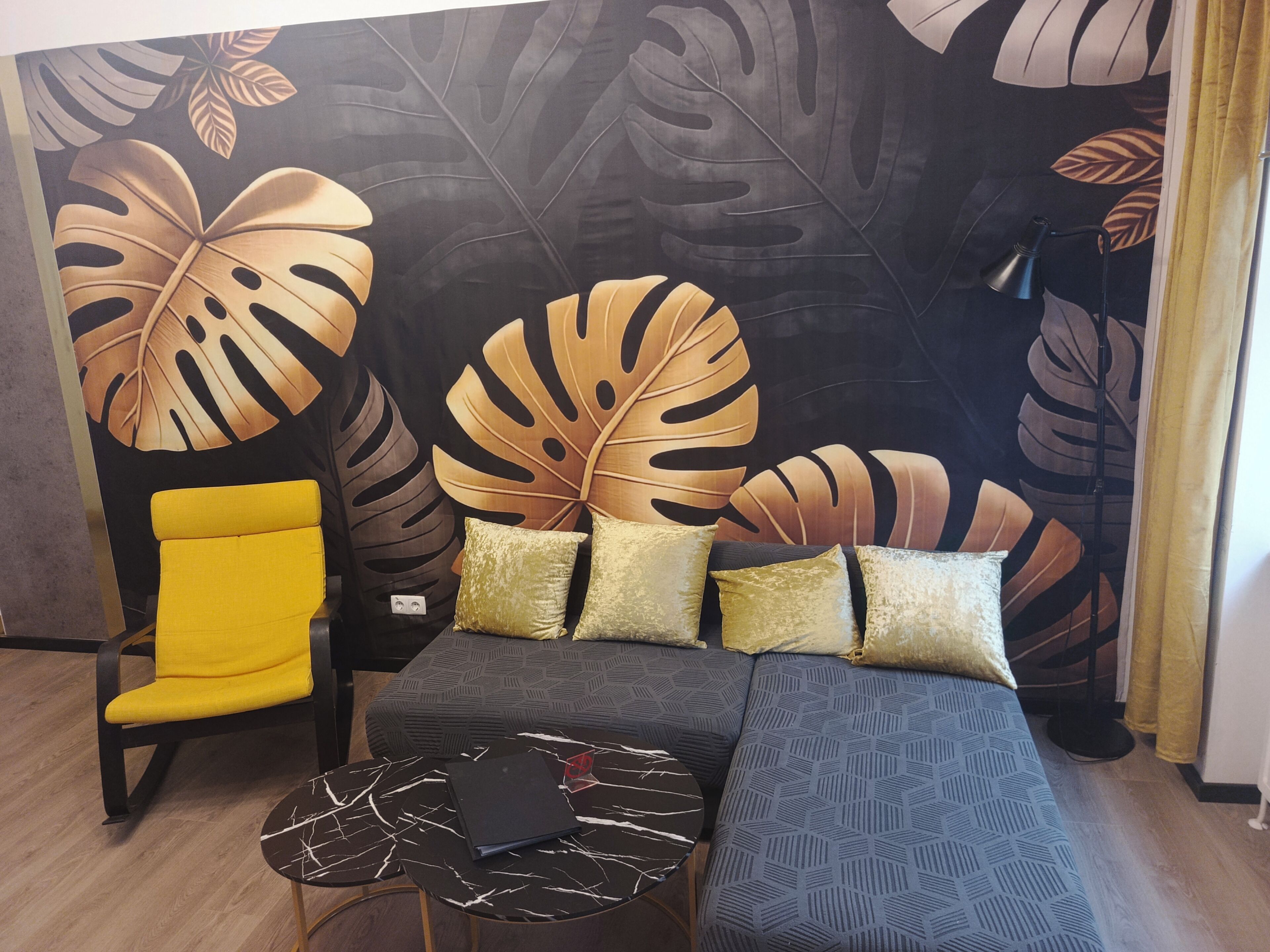 Een kamer met een bed, een gele stoel, een kleine tafel en een groot wandpaneel met bladmotieven.