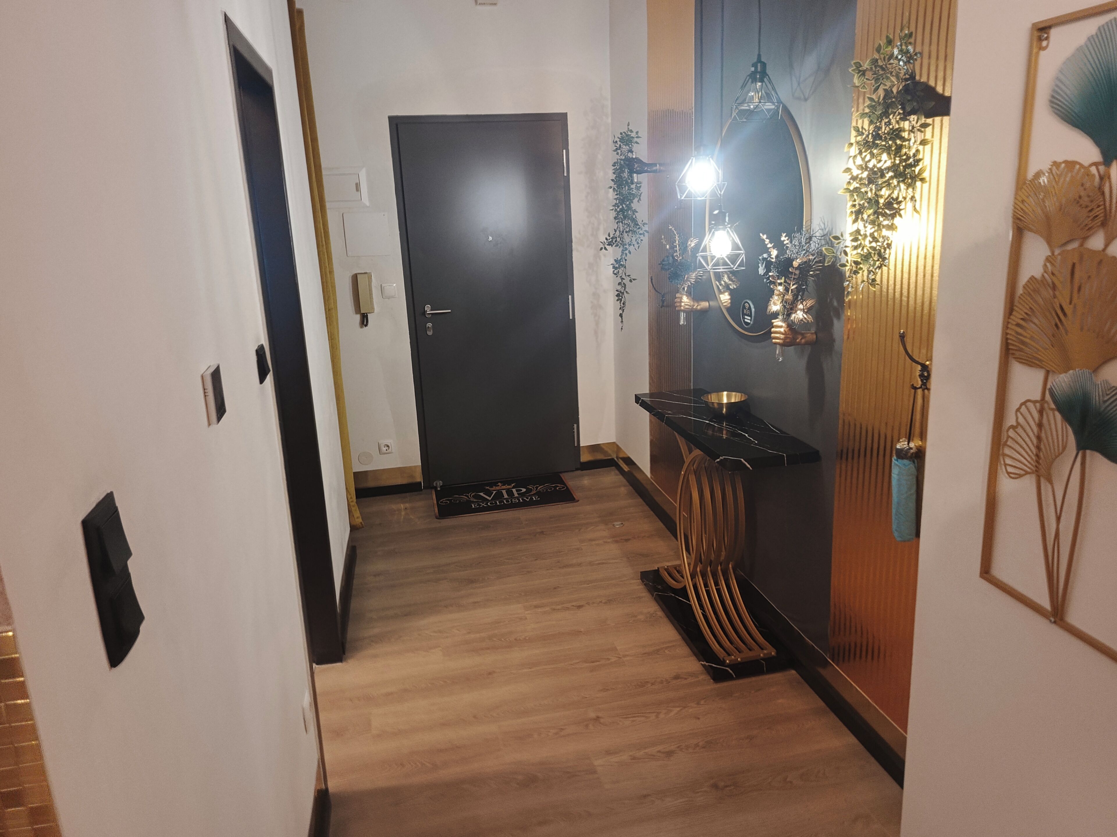En hall med trägolv, en svart dörr, ett litet bord med en skål och en väggmonterad lampa.
