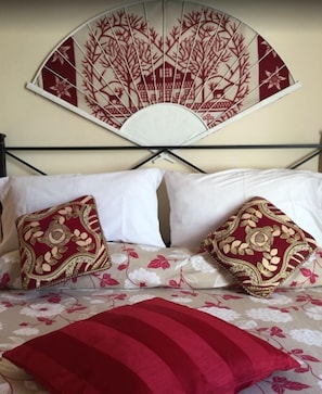 Un lit avec une literie à motifs floraux, deux coussins décoratifs et un ventilateur fixé au mur, orné d’un motif d’arbre.