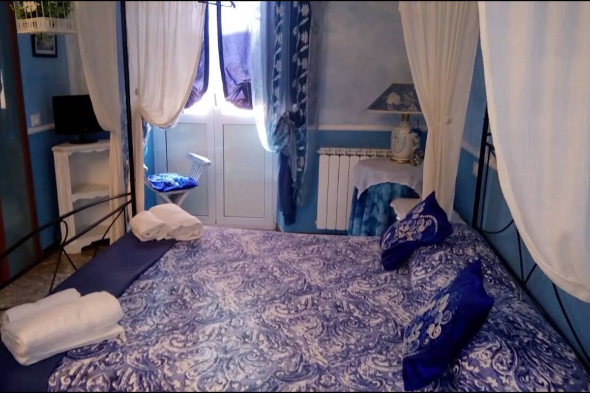 Un dormitorio con cama, cortinas blancas, una ventana, una mesita de noche y una lámpara.
