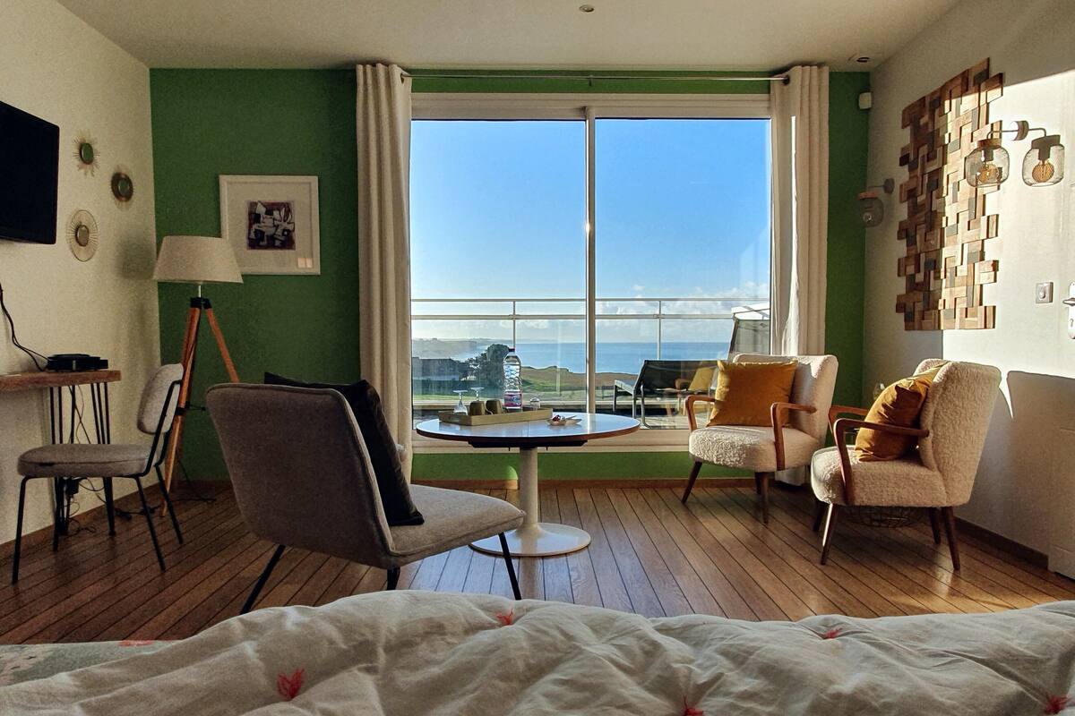 Habitación de hotel con un ventanal grande, una mesa redonda, dos sillones, una cama y un escritorio con lámpara.
