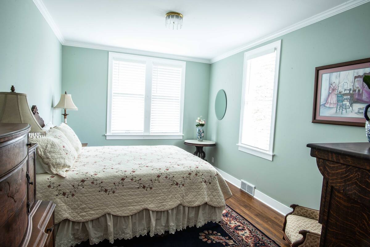 A bedroom with a bed, dresser, lamp, windows, and a painting on the wall.