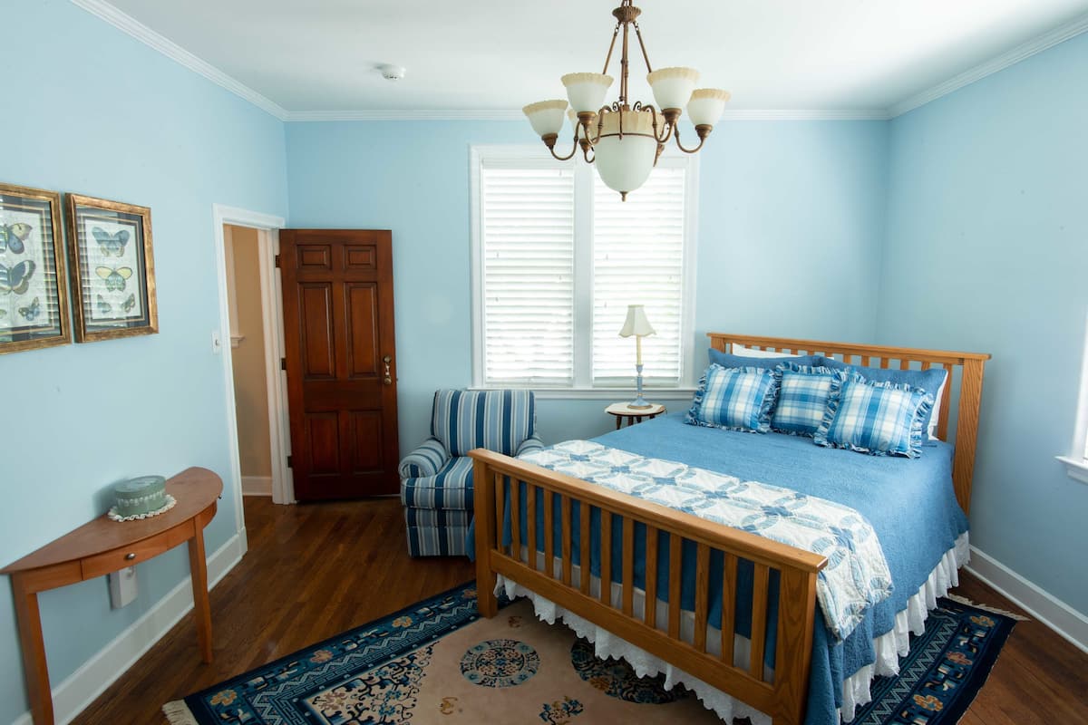 A bedroom with a wooden bed, blue bedding, a chandelier, a window with blinds, a chair, and a rug.