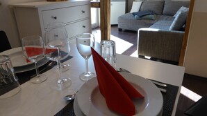 Uma área de refeições com uma mesa posta para dois, incluindo copos de vinho, pratos e talheres. Um guardanapo vermelho está dobrado no prato.