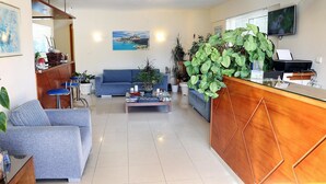 Una sala de estar moderna con un sofá azul, una mesa de centro y un mostrador de recepción de madera. Hay varias plantas en macetas y un cuadro en la pared.