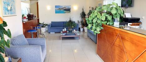 A modern living room with a blue sofa, a coffee table, and a wooden reception desk. There are several potted plants and a painting on the wall.