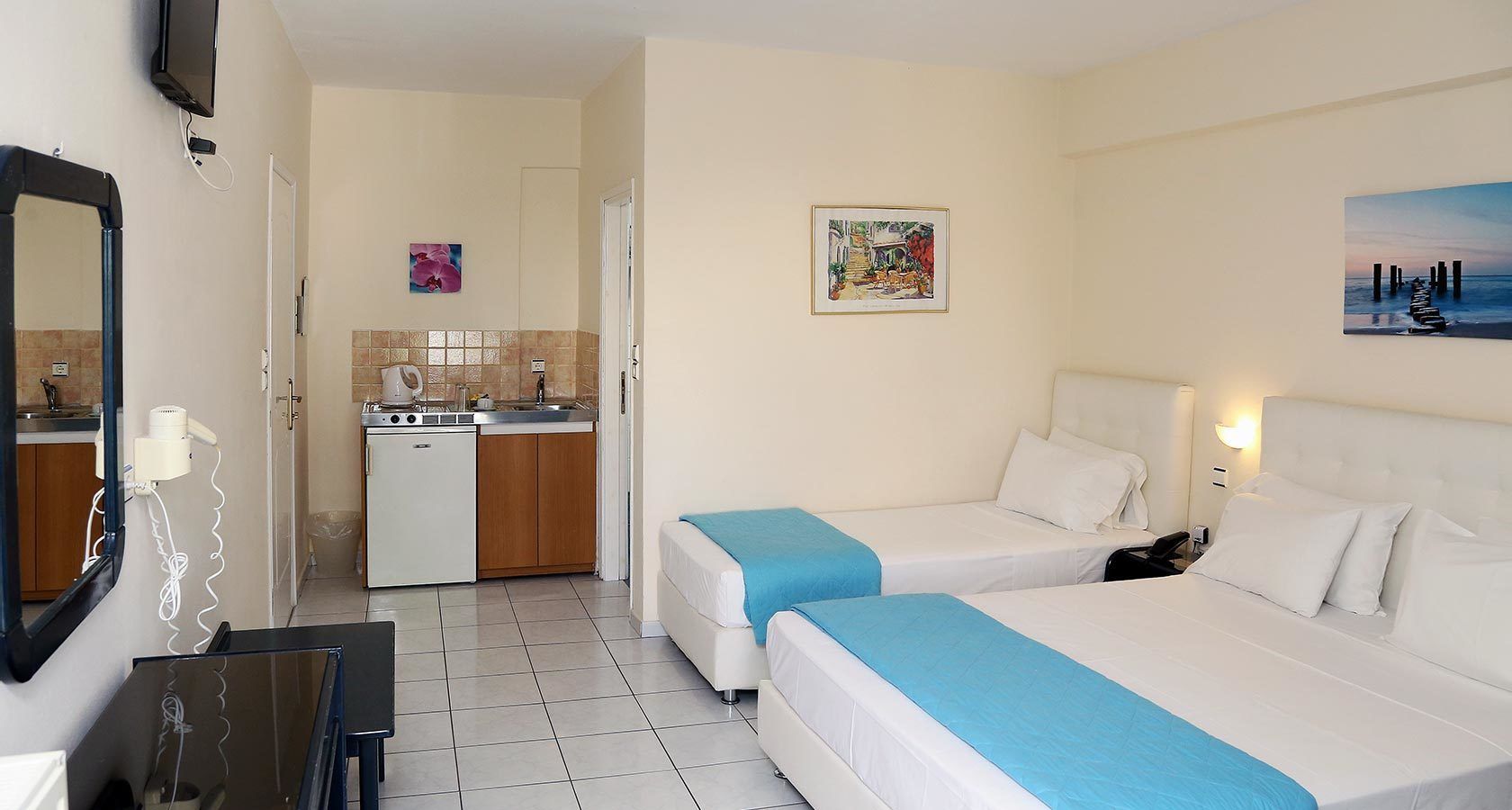 Habitación de hotel con dos camas, un escritorio pequeño, un espejo, un televisor, una zona de cocina y obras de arte en las paredes.