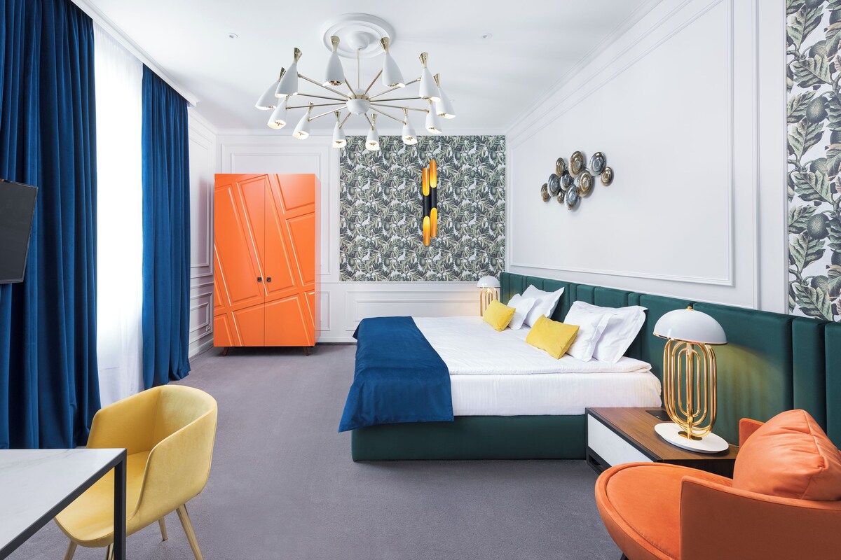 A hotel room with a bed, a desk, a chair, a TV, a chandelier, and a wall with a leaf pattern.