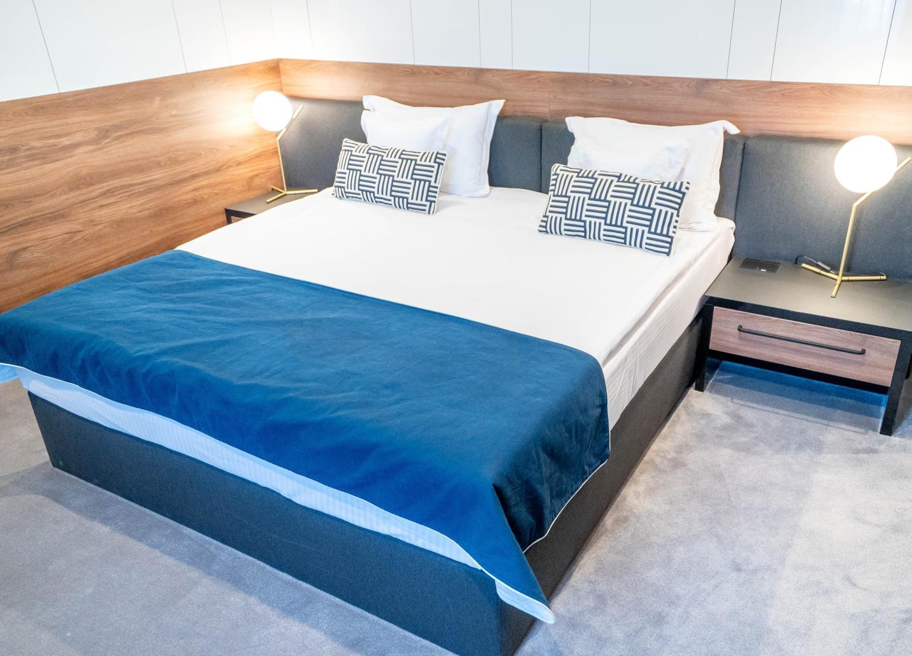 A modern hotel room with a large bed, two bedside tables with lamps, and a built-in bench at the foot of the bed.