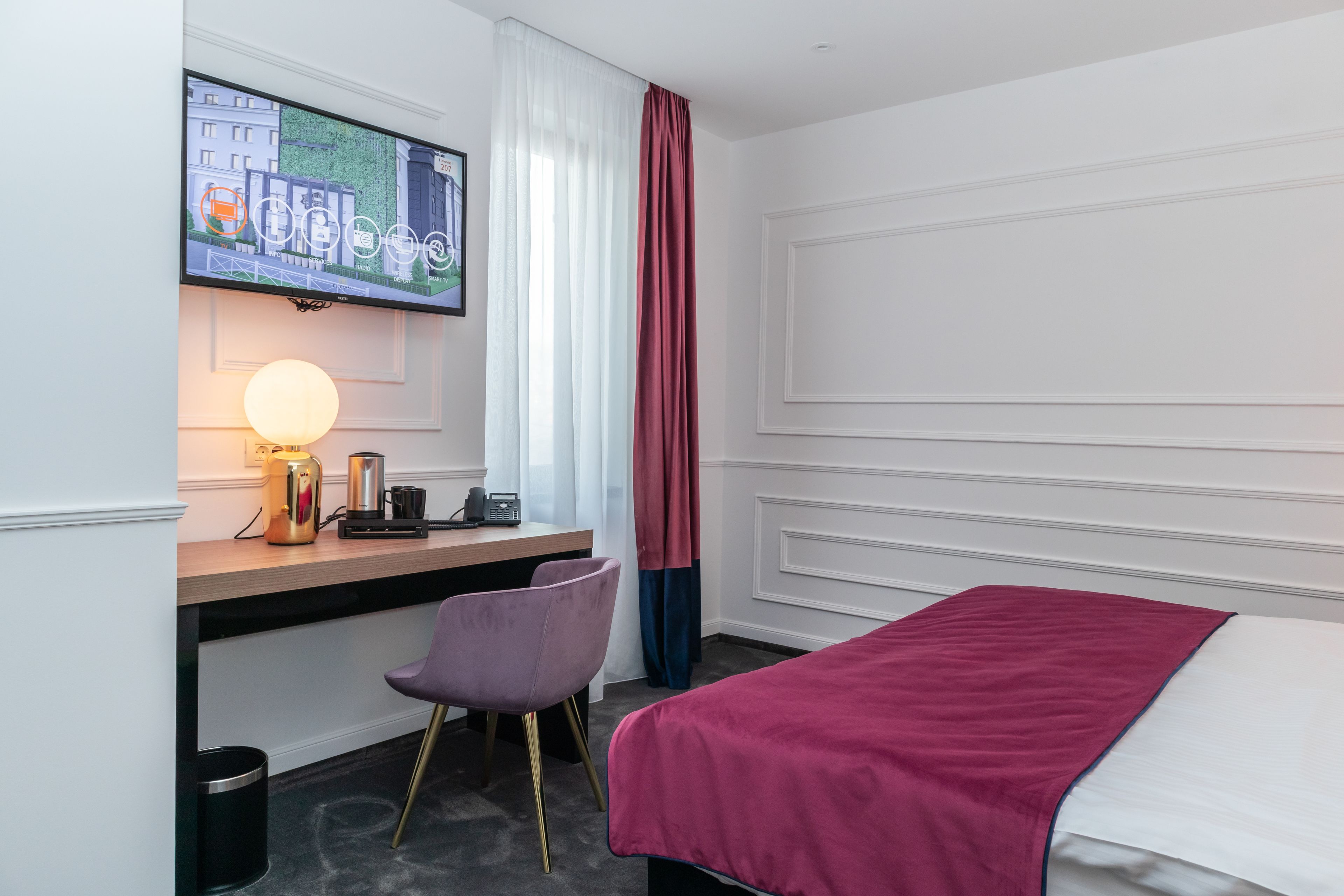 A hotel room with a bed, a desk with a computer, a chair, a TV mounted on the wall, and a window with curtains.