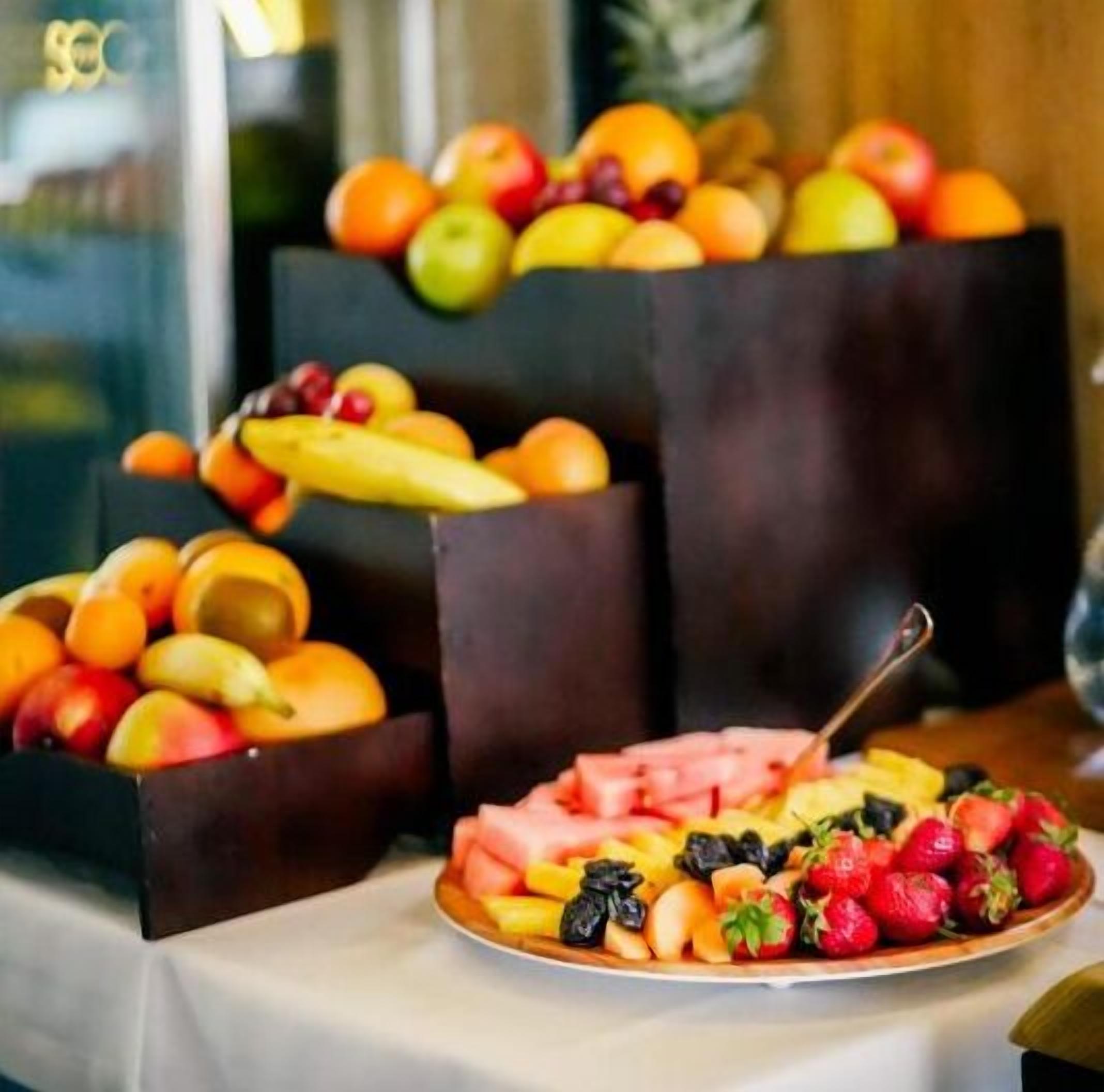 A table with a fruit arrangement including apples, oranges, and a plate with strawberries and other fruits.