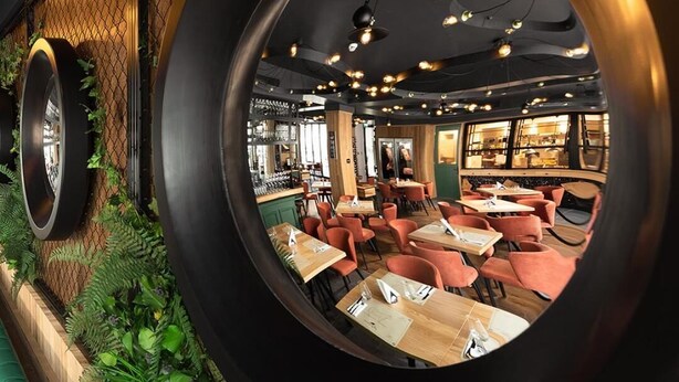 Un restaurante con un ventanal circular, plantas verdes y mesas y sillas de madera.