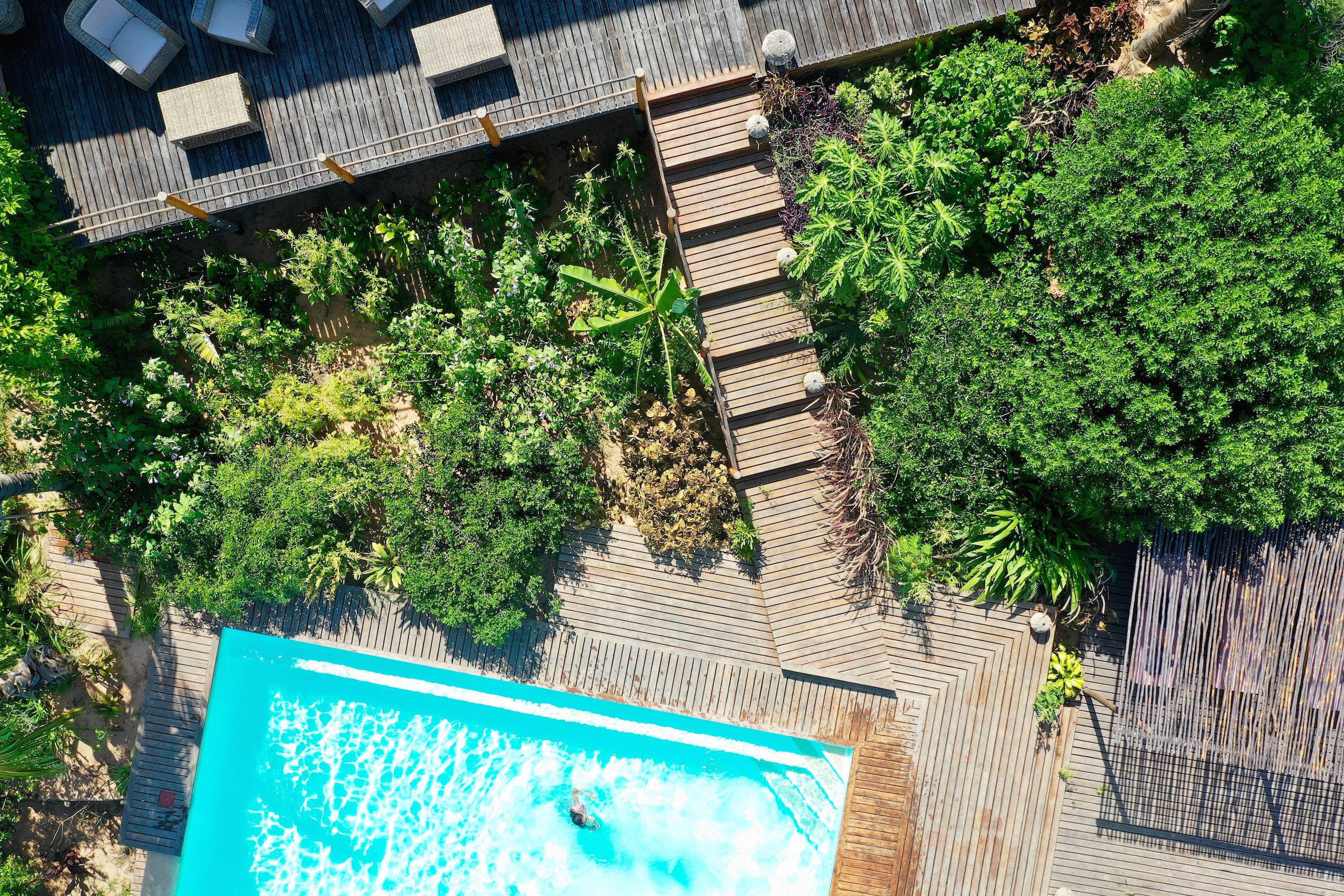 An aerial view of a rooftop pool area with a wooden deck, surrounded by lush greenery and a fenced area.