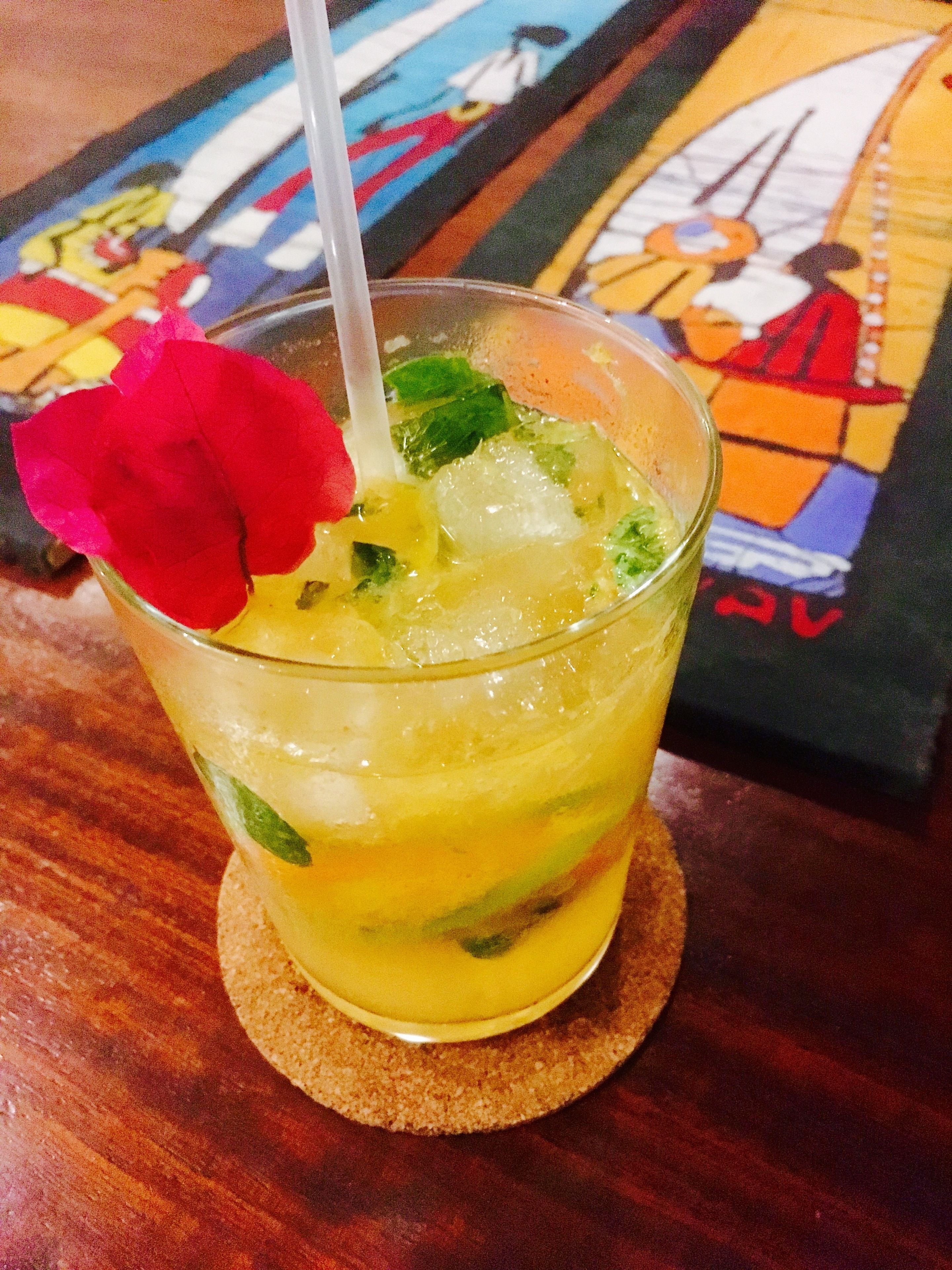 A glass of yellow beverage with a red flower garnish, ice cubes, and mint leaves, placed on a coaster.