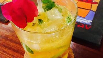 Un vaso de bebida amarilla con una flor roja como adorno, cubitos de hielo y hojas de menta, sobre un posavasos.