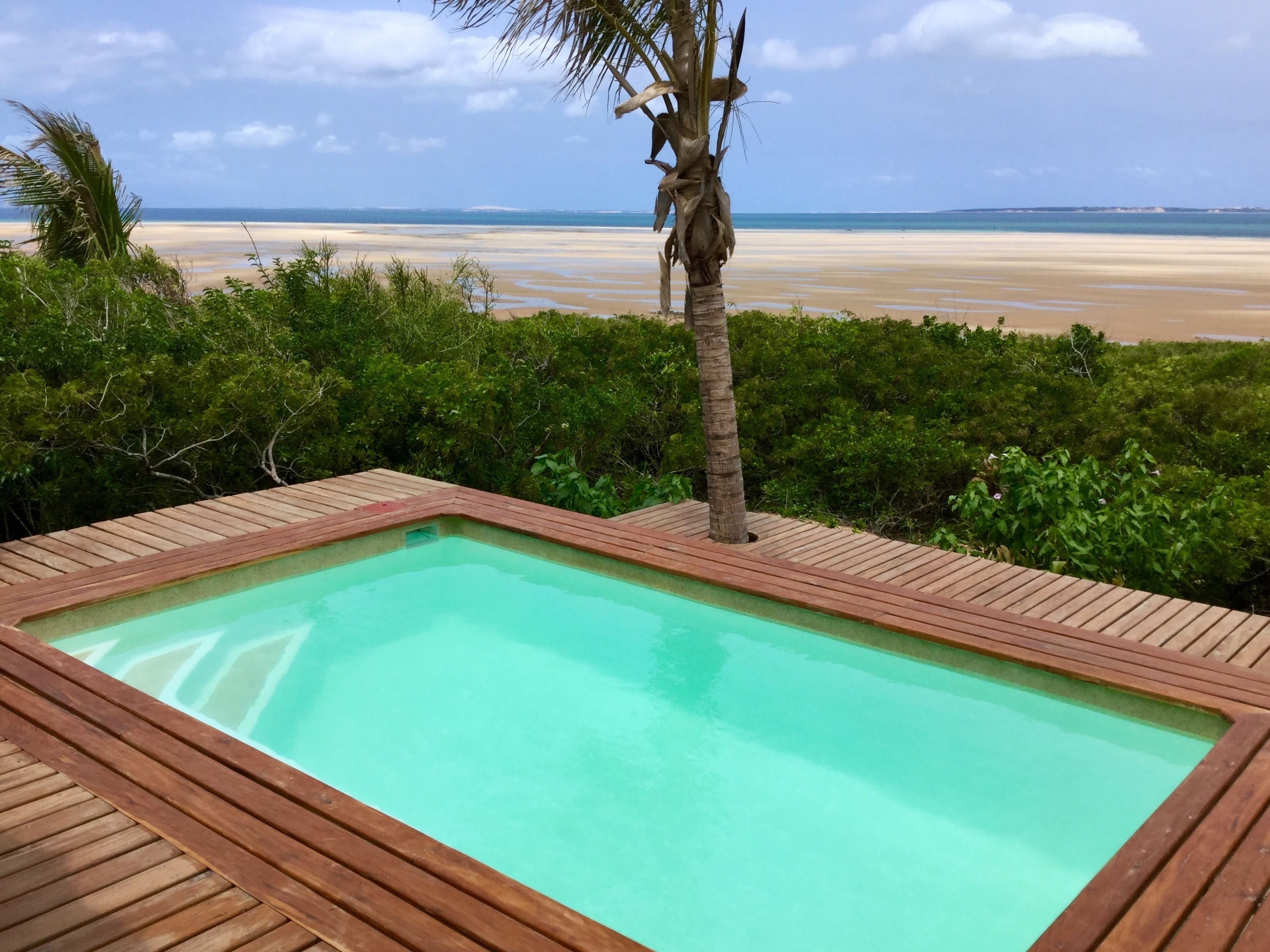 A rectangular pool with a wooden deck, surrounded by lush greenery and a view of a beach and ocean.