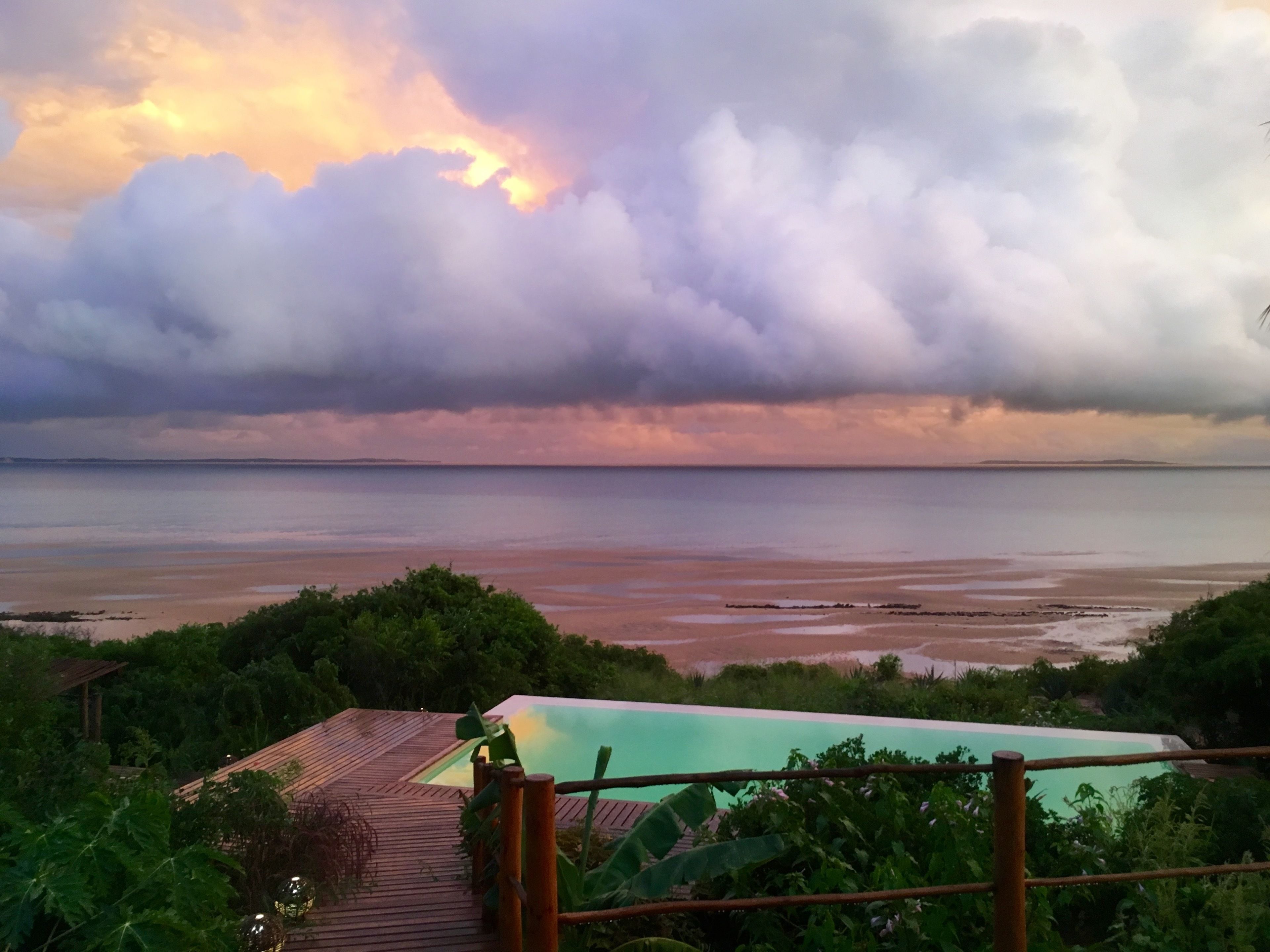 A view of a beach with a wooden deck and a pool, surrounded by greenery.