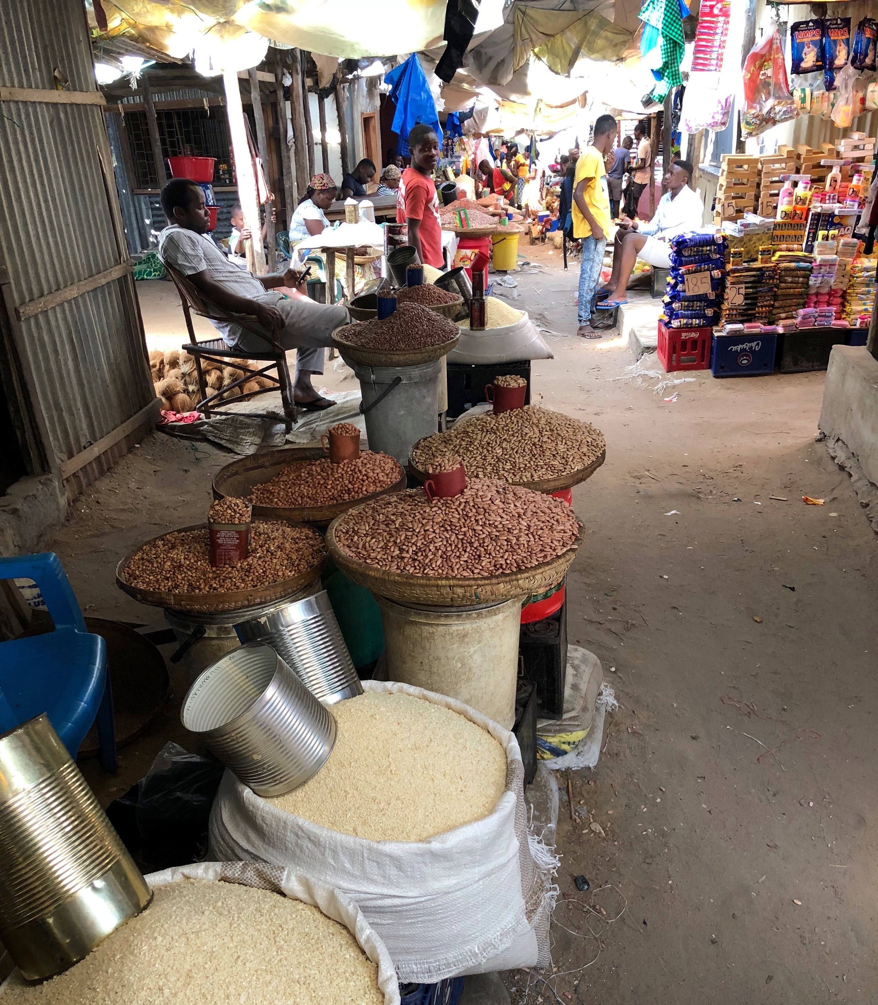 Un mercado con grandes sacos de granos y frijoles, un hombre sentado en una silla, y diversos productos expuestos al fondo.