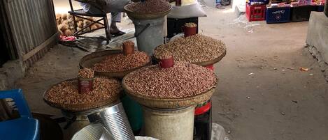 Un mercado con grandes sacos de granos y frijoles, un hombre sentado en una silla, y diversos productos expuestos al fondo.