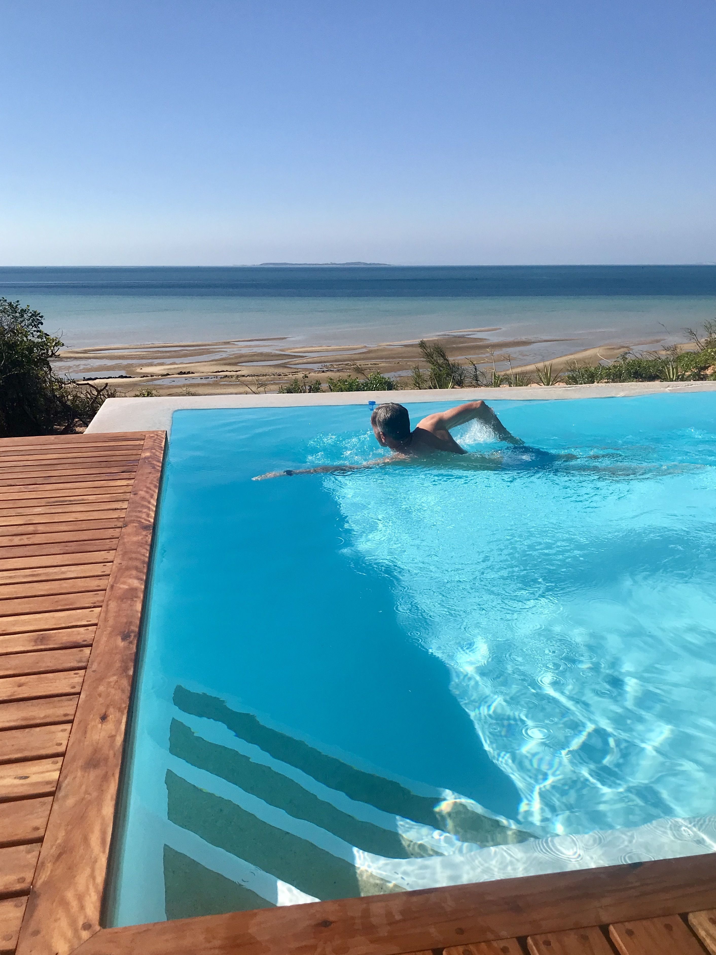 Una persona nada en una piscina azul cristalina con una cubierta de madera que la rodea, con vista a una playa y al océano.