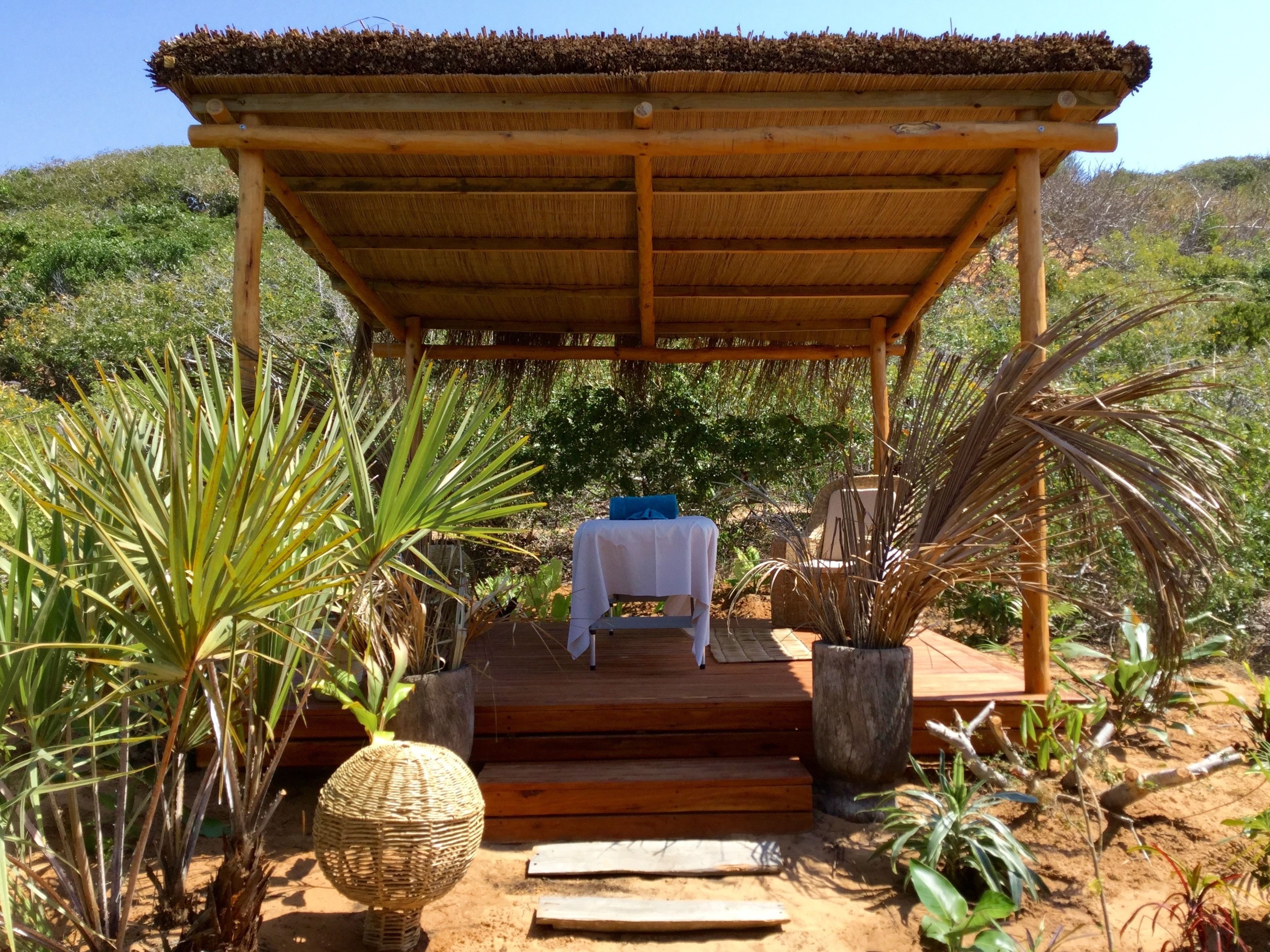 A wooden gazebo with a thatched roof, a covered seating area with a table and chairs, surrounded by various plants and a view of a hilly landscape.