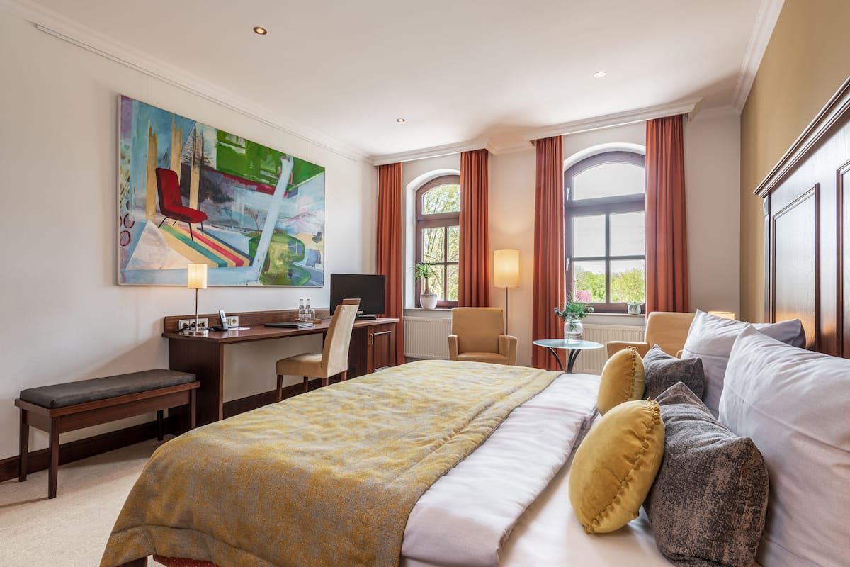 Ein Hotelzimmer mit einem großen Bett, einem Schreibtisch mit Computer, einem Stuhl und einem Fenster mit Vorhängen.