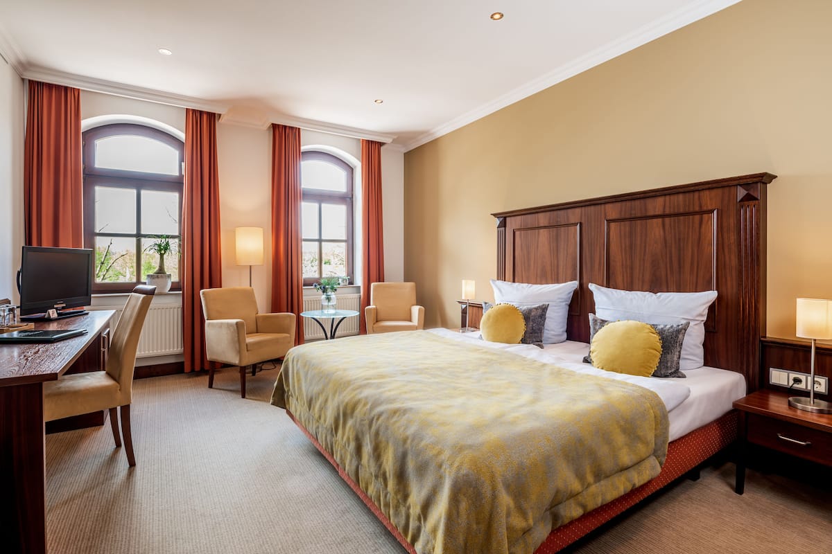 Ein Hotelzimmer mit einem großen Bett, einem Schreibtisch mit Computer, zwei Stühlen, einem kleinen Tisch mit Lampe und einem Fenster mit Vorhängen.