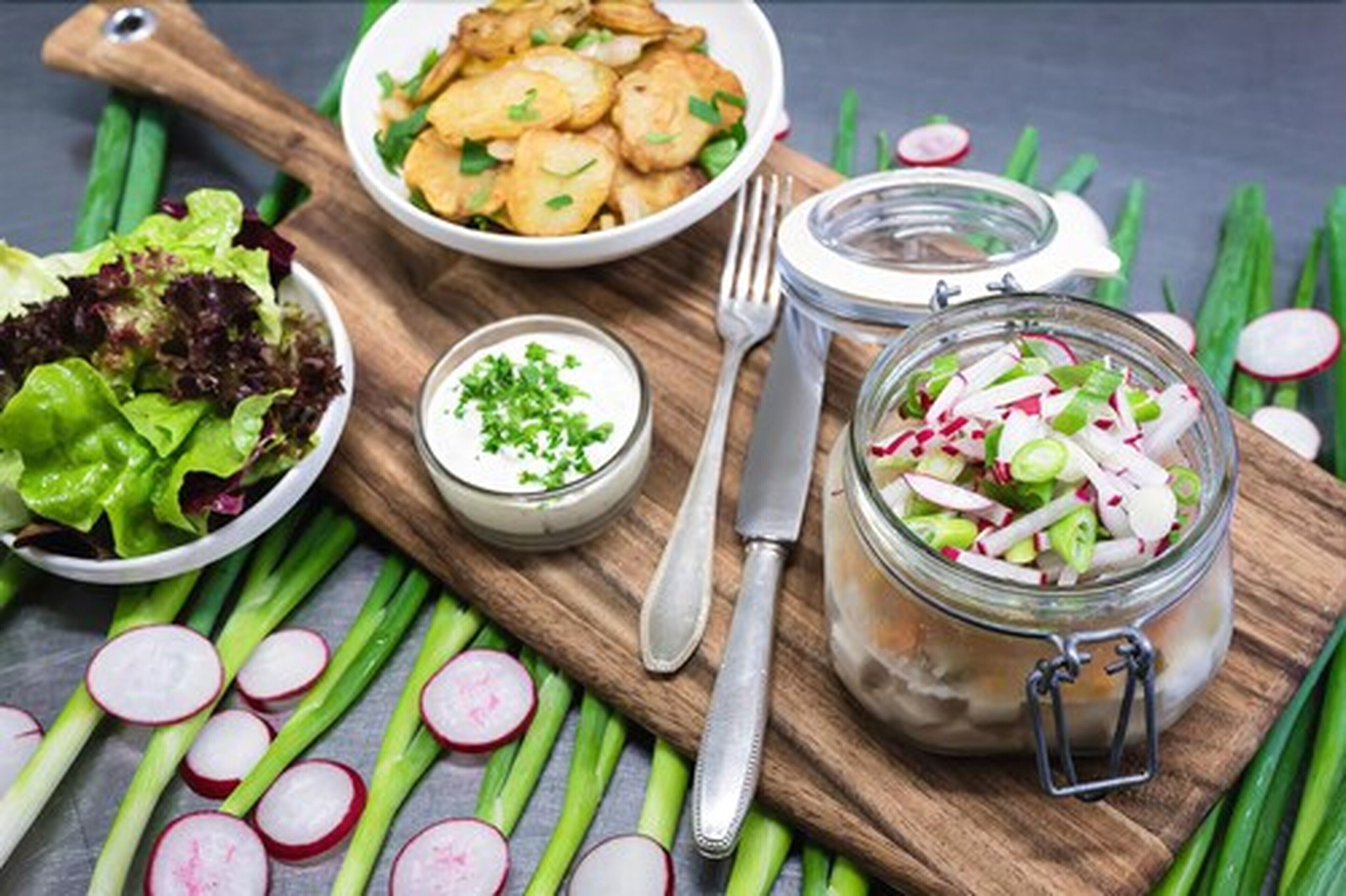 Una mesa de madera con una ensalada, un tazón de papas fritas, un frasco de vegetales encurtidos y un tenedor.