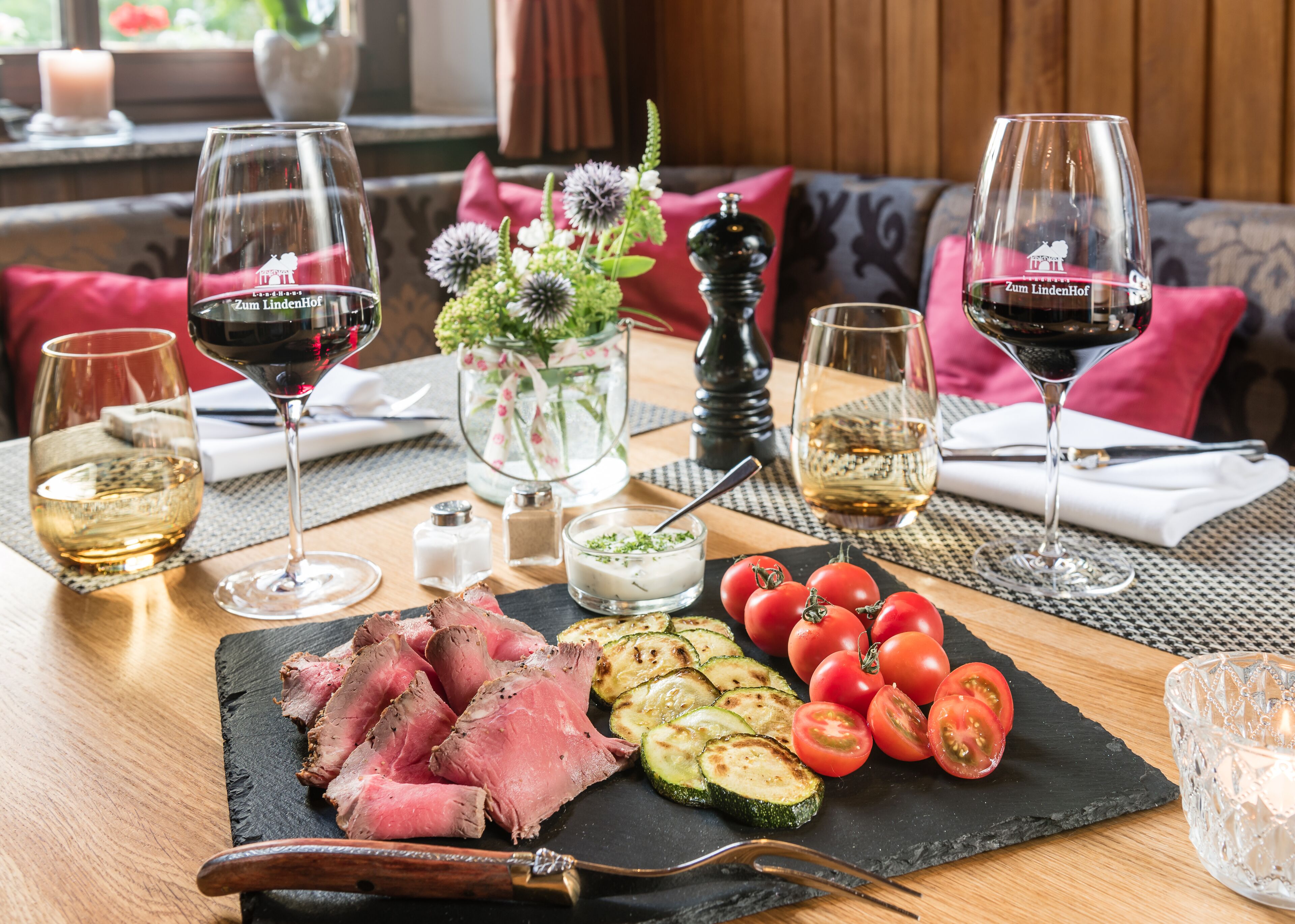 A dining table with a variety of food items, including sliced meat, tomatoes, and grilled vegetables, accompanied by glasses of red and white wine.