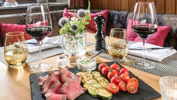 A dining table with a variety of food items, including sliced meat, tomatoes, and grilled vegetables, accompanied by glasses of red and white wine.