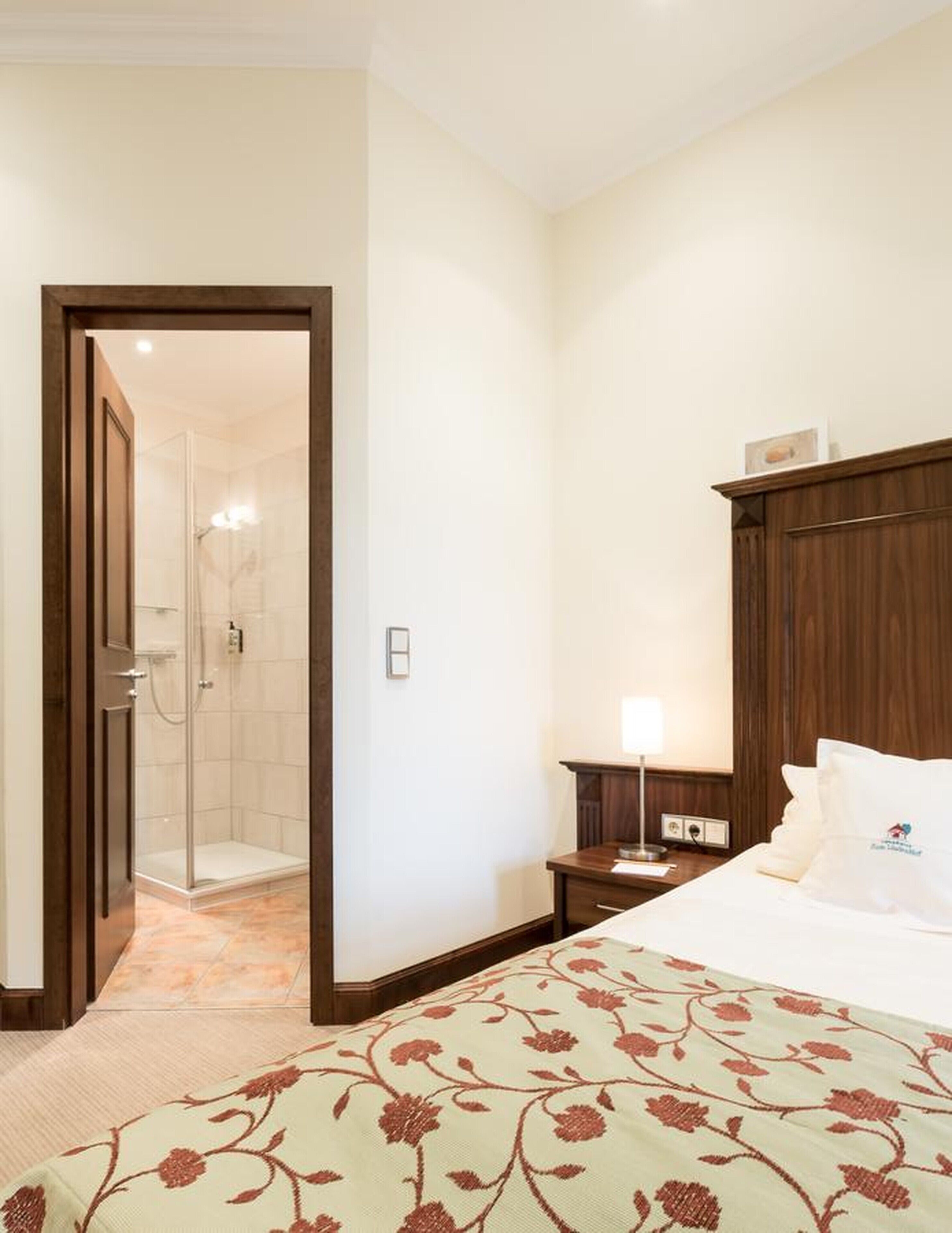 A hotel room with a bed, a wooden headboard, a nightstand, a lamp, and a shower area.
