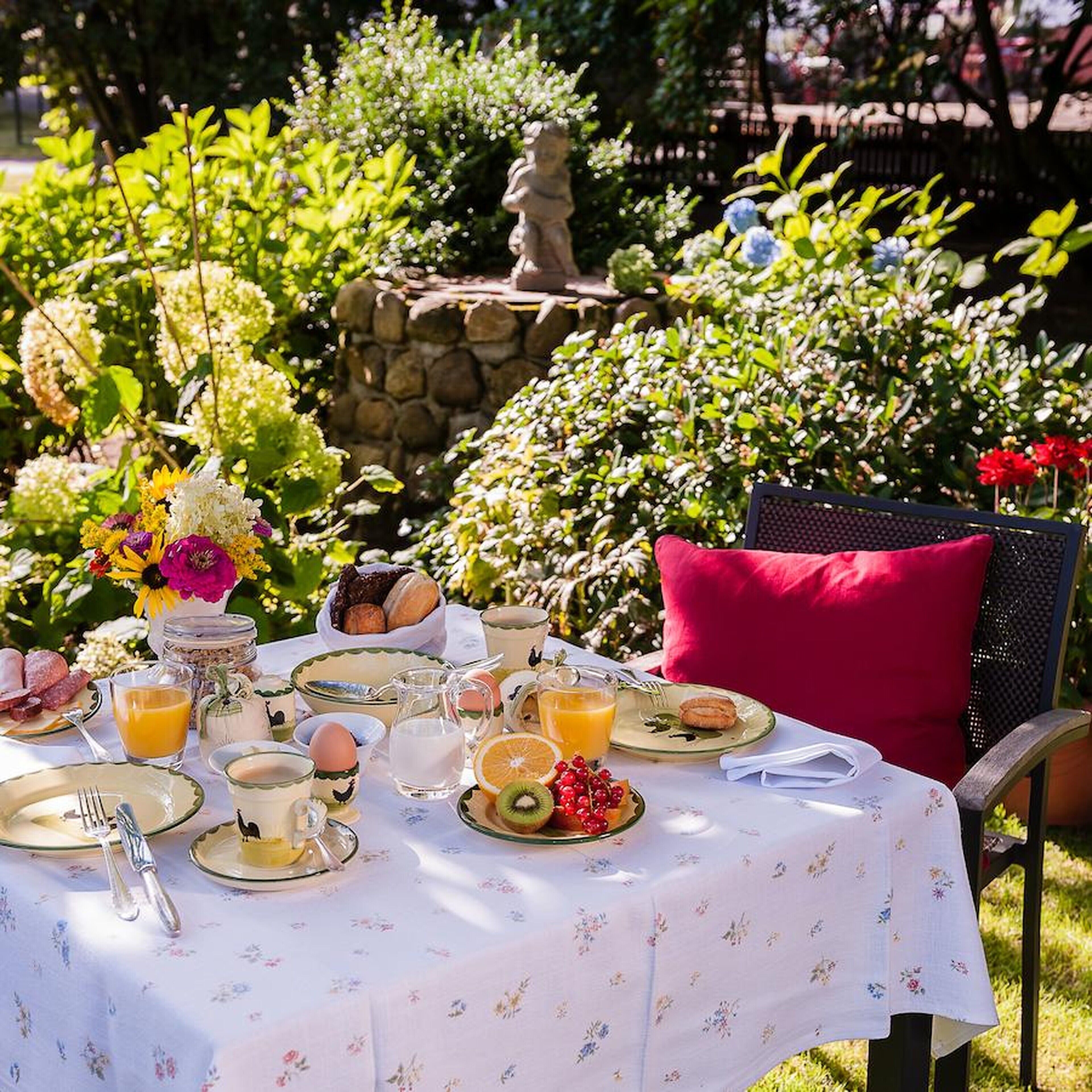 An outdoor dining table set with breakfast items, surrounded by lush greenery and a statue.