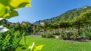 A lush green garden with a clear sky, a mountain in the background, and traditional buildings. - Gartenheim B&B Tramin (Termeno Sulla Strada del Vino)