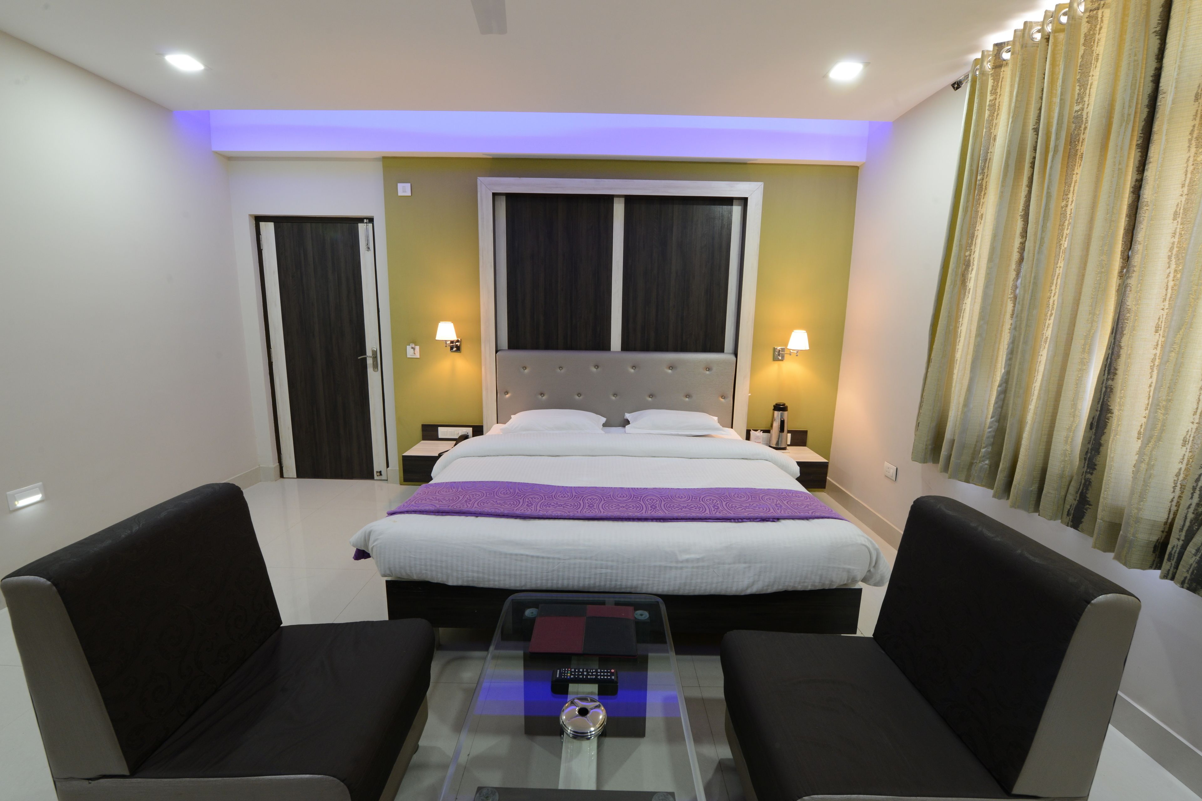 A hotel room with a bed, two bedside tables with lamps, a glass coffee table, and two armchairs.