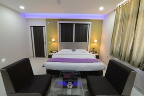 A hotel room with a bed, two bedside tables with lamps, a glass coffee table, and two armchairs. - Hotel Heeralal (Bikaner)