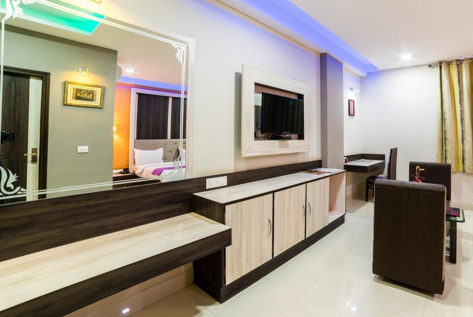 A modern hotel room with a large mirror, a TV mounted on the wall, a wooden bench, and a small table.