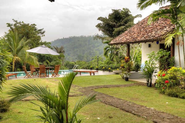 A pool area with a table and chairs, surrounded by lush greenery and a mountain backdrop.