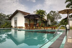 A swimming pool with a patio area, including wooden chairs and a table, surrounded by lush greenery and a building with a tiled roof. - Omatta Villa (Agalawatta)