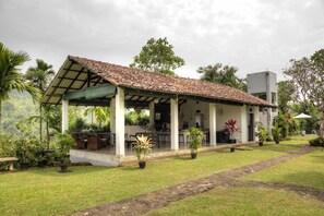 A covered outdoor area with a tiled roof, surrounded by potted plants and a grassy lawn. - Omatta Villa (Agalawatta)