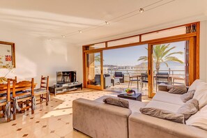 A spacious living room with a sofa, a coffee table, and a TV. There is a balcony with a view of the sea and palm trees. - Emma Beachfront Apartment - Apartment with seaview (Puerto Pollensa)
