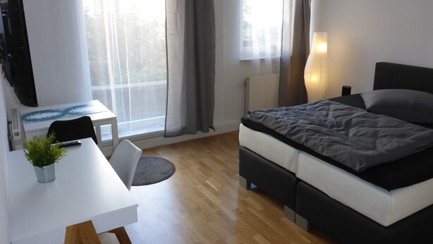 A modern bedroom with a bed, bedside table, lamp, and a desk with a chair and a plant.
