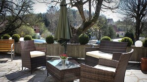 An outdoor seating area with wicker furniture and a glass table, surrounded by stone flooring and greenery.