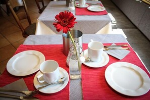 Una mesa de comedor dispuesta con platos blancos, tazas y cubiertos, un florero con una flor roja y una botella de vidrio transparente.